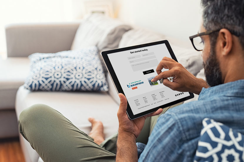 Man hand working with tablet and blueprint on architectural project at home while relaxing on couch. Back view of latin architect reading plan layout on digital tablet.